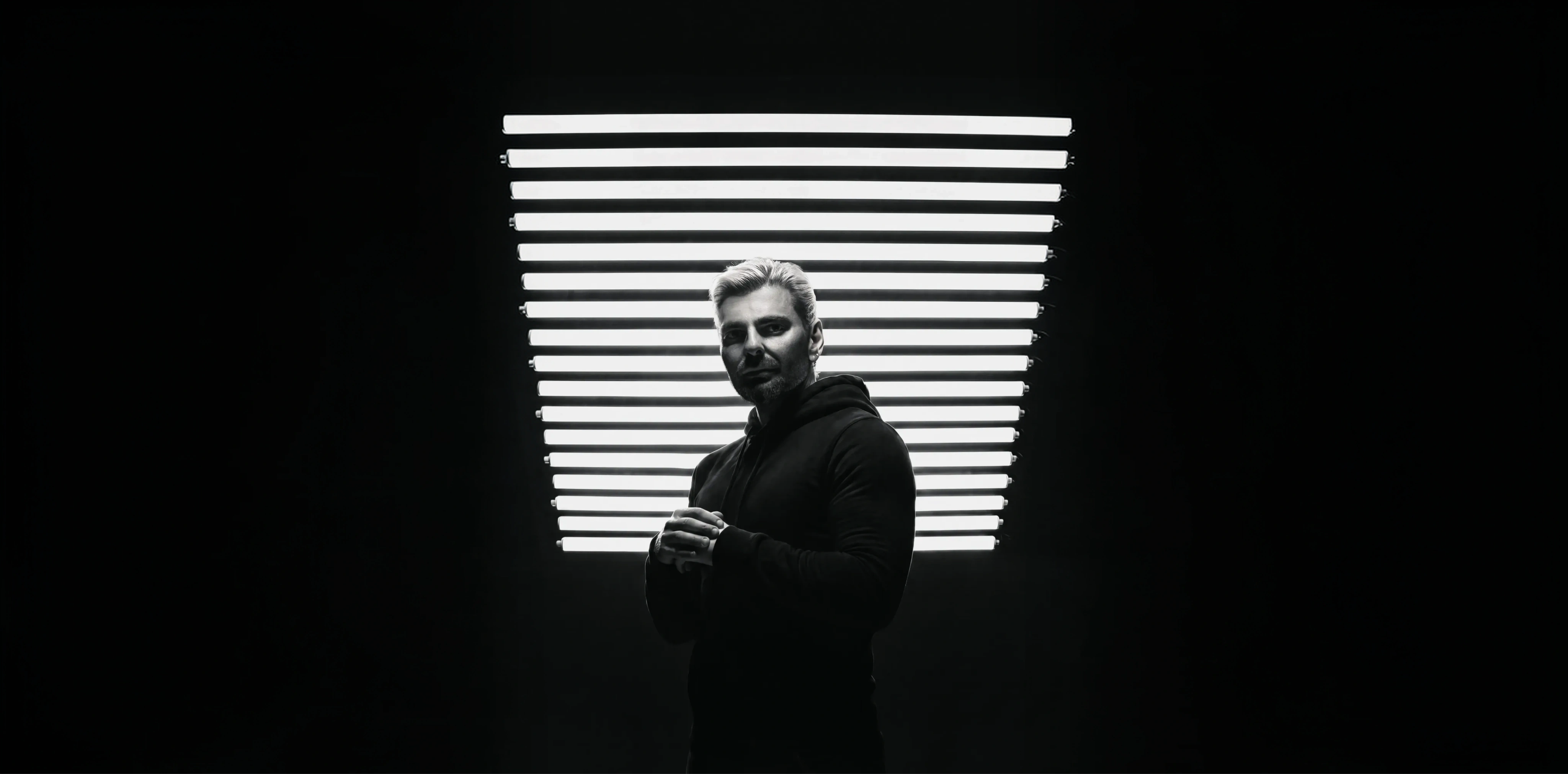 Lucas Rosa, em preto e branco, diante de uma parede de tubos de luz